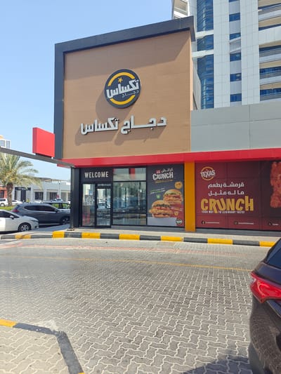 The image shows the exterior of a Texas Chicken restaurant located in Sharjah, United Arab Emirates. The restaurant has a prominent sign with Arabic text. A large glass door with "WELCOME" written above it is visible, along with large advertisements on the walls displaying food items and the restaurant's branding. Several cars are parked in front of the restaurant on a paved lot. The sky is clear and blue, and a modern building is visible in the background. The overall scene depicts a commercial area in a city.