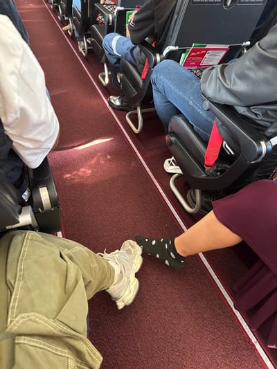 An overhead shot on what appears to be an airplane. Rows of seats are visible with passengers seated. One person wears black and white polka dot socks, another khaki pants and sneakers. A carry-on bag sits on the floor. The carpet is red with thin white stripes. Magazines are in the seat pockets. The overall composition suggests a cramped or tight space.