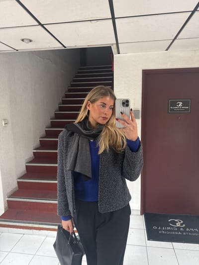 A blonde female adult is captured in a mirror selfie within the interior hallway of a building located at 8, rue Laroque, Aix-en-Provence, France. She is seen from the waist up, wearing a blue long-sleeved top, a dark gray textured jacket, a dark gray scarf, and black trousers. Her right hand holds a light gray ribbed smartphone with a visible camera array, used for the selfie, and her fingernails are painted dark red. A black handbag is held in her left hand. The environment consists of a light-tiled floor, a light-colored stuccoed wall with a small rectangular fixture, and a dark red door on the right. The door features a mirrored sign reading 'SAPPA & CIRILLO AVOCATS ASSOCIES' and an adjacent access control panel. A mirrored floor mat with the same text is positioned below the door. To the left, a staircase with red treads and gray risers ascends into a dimly lit upper level. The ceiling is a grid-panel type with a single recessed light fixture visible.