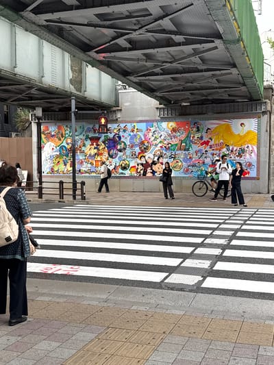 The image shows a vibrant, large mural depicting various anime characters and scenes, stretching along a building wall under an elevated train track structure in Shinjuku, Japan. In the foreground, a pedestrian crossing with white stripes dominates the scene. Several people are visible: a woman in a patterned top and dark pants with a beige backpack stands on the left, facing away from the camera. To the right, near the mural, a man in a white shirt and dark pants with a backpack walks across the street, while a woman in a black outfit stands near him, looking to the side. Another woman in black pants and a black top stands to the left of the mural, with a bicycle parked beside her. The mural itself is colorful and detailed, featuring figures reminiscent of classic Japanese animation. The environment appears urban, with concrete and metal structures overhead, suggesting the presence of public transportation and city infrastructure. The overall scene captures a moment of everyday life in a bustling city, with the striking mural adding a unique artistic element.