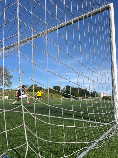 A soccer field at Champigny-sur-Marne, France, is viewed through the netting of a goal. Two soccer players are in motion on the field. One player wears a yellow shirt and dark shorts, while the other wears a dark shirt and white shorts with red shoes. The sky is blue with light clouds. Trees and buildings are visible in the background.