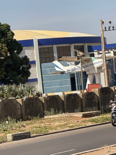 The image depicts a daytime scene in Basawa, Nigeria, featuring a static aircraft display in front of a modern building. A white, single-engine, high-wing aircraft, registered "5N-AZR," is prominently mounted on a tall, angular white pedestal. The aircraft has blue accents on its tail. Behind the aircraft is a large building with a light yellow corrugated metal roof, blue trim, and a facade composed of dark windows, blue horizontal panels, and white lattice sections. To the left of the building, a large tree with dark green foliage is visible. In the foreground, a paved asphalt road runs horizontally, separated from the property by a low, dark gray concrete barrier wall. Between the road and the barrier, sparse green and dry vegetation covers the ground. Behind the barrier, closer to the building, are several spiky, light green plants. On the far right, the front wheel and handlebars of a dark-colored motorcycle are visible on the road, with a partially obscured rider. A utility pole with multiple cross-arms and wires stands on the right side of the frame. The sky is clear and light blue.