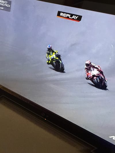 The image shows a replay of a motorcycle race on a screen. Two motorcycles are visible, one red and white and the other yellow and black, racing on a gray track. The red and white motorcycle is in the lead, with the yellow and black motorcycle following closely behind. The "REPLAY" logo is visible on the left side of the screen, indicating that the footage is a replay. The "MotoGP GRAND PRIX" text is also visible at the bottom left of the screen. The scene appears to be a sporting event, likely a professional motorcycle race. There are no clear indicators of time of day or weather conditions. The image is a photograph of a screen, and the overall setting is indoors, as indicated by the surrounding dark frame and the textured background on the right.