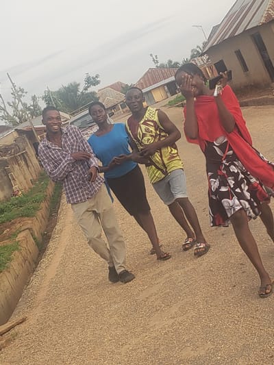 The image depicts four young individuals outdoors on a dirt road, likely in the city of Gumel, Nigeria. The individuals are positioned in a line, with their bodies angled towards the viewer. The person on the far left is a male wearing a checkered shirt and light-colored pants. Next to him is a female in a blue top and dark skirt. To her right is a male in a sleeveless patterned shirt and shorts. The person on the far right is a female wearing a red wrap over a floral print dress. She appears to be holding a phone in her right hand and covering her face with her left, possibly in a gesture of surprise or shyness. The background shows a collection of buildings with corrugated metal roofs, characteristic of residential structures in the region. The ground is a mix of dirt and small gravel. The sky is overcast, suggesting a cloudy day.