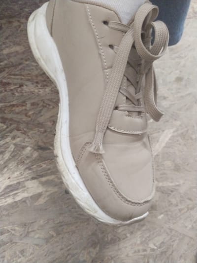The image shows a close-up of a light brown lace-up sneaker with a white sole, resting on a textured wooden surface. The sneaker appears to be slightly worn, with some scuff marks visible on the sole. The laces are tied in a bow, and a white sock is visible peeking out from the top of the shoe. The background is a blurry, light-colored wooden surface with a visible grain pattern. The scene is captured from a slightly elevated angle, focusing on the shoe. The location is identified as Edineț, Moldova.