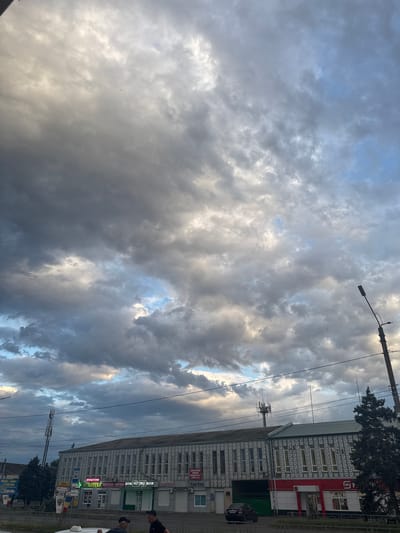 A frontal, ground-level, vertical shot captures a commercial building in Yegorlykskaya, Russia, under a dramatic sky filled with heavy, grey clouds, interspersed with patches of blue. The building, which appears to be a single-story structure with a two-story section, features a repetitive patterned facade. Several businesses are identifiable by their signage, including an "APTEKA" (pharmacy), a "MYASNAYA FERMA" (meat farm), and a store with the logo "M" and the word "МАГНИТ" (Magnit), a popular Russian supermarket chain. A streetlight stands prominently on the right side of the frame, and a telecommunications tower is visible in the distance above the building. In the foreground, a section of a white vehicle is visible, and two men are standing on a sidewalk in front of the building, one wearing a black t-shirt and the other a checkered shirt and a black cap. The overall impression is of a cloudy, possibly late afternoon or early evening scene in a Russian town.