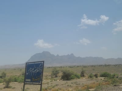A wide shot captures a vast, arid landscape under a hazy, pale blue sky dotted with sparse clouds. The terrain is mostly flat, covered in dry brush, scattered rocks, and a few small, green shrubs and trees. In the background, a range of rugged, dusty mountains stretches across the horizon, their peaks sharp and defined against the pale sky.

In the foreground, on the left side of the image, a blue sign with white Arabic script stands on a metal pole. The script appears to be a directional sign. Below the main text, a white arrow points to the left. The sign is slightly blurred, but the text is clearly visible.

The overall impression is of a remote, undeveloped, and dry environment. The lighting suggests it is daytime, likely mid-morning or afternoon, given the brightness of the sky. The hazy atmosphere and muted colors indicate a hot, dry climate. No people are visible in the frame, and the scene appears still and undisturbed, suggesting a quiet, natural setting. The sign indicates a point of reference or direction within this expansive, rugged landscape, possibly a road or a place of interest.
