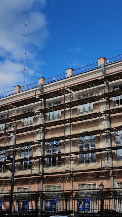 Captured from a low angle, the photograph presents the facade of a multi-storied building in Smolensk, Russia, likely undergoing restoration. Scaffolding obscures much of the building's features, but visible are windows of varying shapes, ornate detailing between floors, and a rooftop with a decorative blue railing. The building's exterior is a light brownish-tan color.

The background showcases a bright blue sky dotted with fluffy white clouds. In the lower-left corner of the frame is a tall antenna. Additional structural metalwork and signage are apparent in the lower-right section of the image.