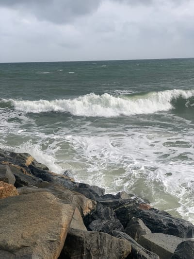 The image depicts a dynamic coastal scene in Ibeju, Nigeria, under an overcast sky. The upper third of the frame shows a uniform grey, cloudy sky meeting the dark green-grey ocean at the horizon line. A large, white-crested wave breaks horizontally across the mid-ground of the water, generating extensive white foam and turbulent water that extends towards the foreground. The lower portion of the frame features a rugged shoreline composed of large, angular rocks, varying in color from dark grey and black to lighter brown and tan. Ocean waves are actively crashing against these rocks, creating white spray and foam where the water meets the land barrier. No individuals or vessels are visible within the frame.