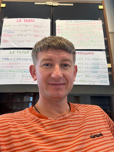 The image shows a medium close-up shot of a man's face and upper torso, taken outdoors. The man is looking directly at the camera with a slight smile. He is wearing an orange and white striped t-shirt with the word "alprausch" embroidered in black on the left side of his chest.

Behind the man, a bulletin board is visible, covered with several handwritten notes in French. These notes appear to be educational materials, possibly for a language class. One section is titled "LA FRÉQUENCE" (Frequency) and includes percentages and phrases related to how often actions occur. Another section is labeled "LA DURÉE" (Duration) and features a timeline with dates and events. A third section is titled "LE FUTUR" (The Future) and discusses verbs in the infinitive and irregular verbs.

The lighting suggests it is daytime, but the overall scene appears to be indoors or in a shaded outdoor area, given the quality of light and the presence of the bulletin board. The background is blurred, but hints of a room with shelving or furniture can be seen. The man's expression is friendly and approachable. The presence of the bulletin board and the educational content strongly suggests a school or learning environment. The word "Bellelay" in the prompt provides a specific location context for this scene.