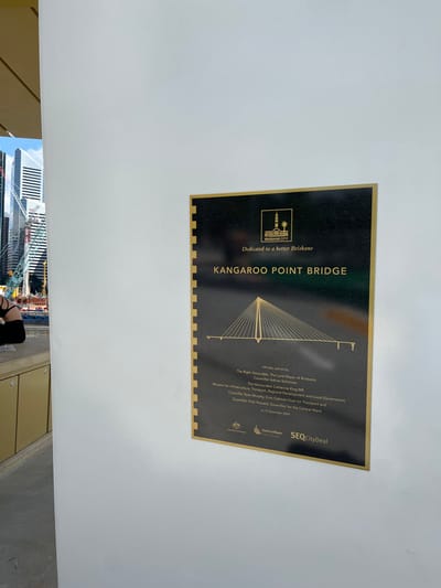 The image displays a dark, rectangular commemorative plaque with gold lettering, oriented vertically against a light background. The plaque is dedicated to the "KANGAROO POINT BRIDGE" and features a stylized drawing of a cable-stayed bridge. Above the bridge name, the text "Dedicated to a better Brisbane" is visible alongside the "BRISBANE CITY" logo, which depicts a building.

The plaque details the official opening, stating it will be "OFFICIALLY opened by" several dignitaries, including "The Right Honourable, The Lord Mayor of Brisbane Councillor Adrian Schrinner," "The Honourable Catherine King MP Minister for Infrastructure, Transport, Regional Development and Local Government," "Councillor Ryan Murphy, Civic Cabinet Chair for Transport and," and "Councillor Vicki Howard, Councillor for the Central Ward." The date specified for this event is "15 December 2024." Additionally, the "SEQ CityDeal" logo and logos for the Queensland and Australian Governments are visible at the bottom right of the plaque.

In the blurred background, modern high-rise buildings and construction equipment, possibly a crane, indicate an urban setting in Brisbane City. A person's feet and lower legs, wearing black open-toed sandals or heels, are visible in the bottom left corner of the original image, partially obscured. The scene appears to be taking place during the daytime under clear weather conditions.
