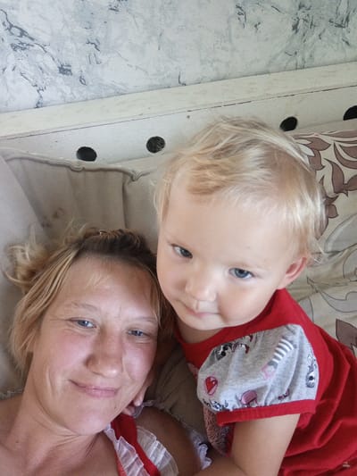 This is a close-up selfie of two individuals, a woman and a young child, lying down. The woman, with blonde hair and blue eyes, is in the foreground, smiling at the camera. The child, also with blonde hair and blue eyes, is slightly behind her and looking towards the camera. Both appear relaxed and happy. They are lying on a surface that looks like a bed or sofa with patterned cushions and a white wooden headboard. The background shows a textured wall with a grey and white marbled pattern. The lighting suggests it is daytime. The child is wearing a red shirt with grey sleeves that have a pattern of cartoon animals and hearts. The woman is wearing a red and white ruffled garment. The image was taken in Buraly, Russia.