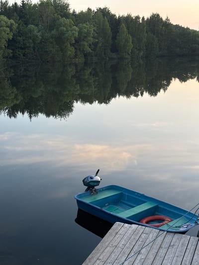 A blue boat with a small outboard motor is docked at a wooden pier on a calm body of water. The boat is empty and contains a red life preserver. The opposite shore is lined with a dense forest of green trees, whose reflections are visible on the water's surface. The sky above is a pale, muted color, suggesting either dawn or dusk. The image appears to have been taken in Kamysh, Russia.