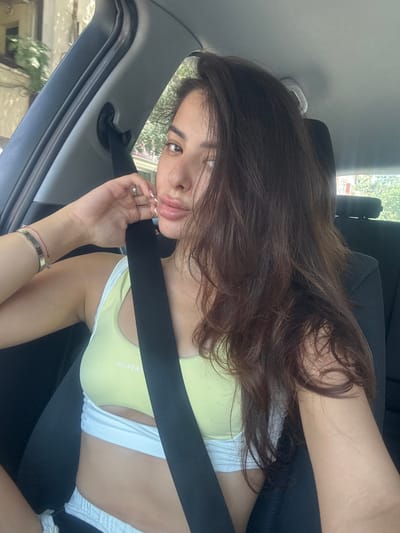 This media file is a portrait of a young woman sitting in the passenger seat of a car. She has long, dark brown hair that partially obscures her face. She is wearing a light yellow sports bra with white trim and white shorts. A black seatbelt crosses diagonally across her chest, secured by the car's seatbelt buckle. Her left hand, adorned with a ring, is raised to her lips, and she wears a bracelet on her left wrist.
The setting appears to be a bright, sunny day, suggested by the light filtering into the car. Outside the car window, greenery and trees are visible, indicating an outdoor location, possibly a suburban or residential area. The interior of the car, including the car door frame and seatbelt, is visible. The woman's expression is casual and relaxed, with a slight smile. The visible text on her sports bra reads "MUSCULAR." The image is taken from a low angle, looking up at the woman. The overall context suggests a candid moment captured while traveling.