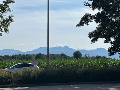 The image captures a serene daytime scene in Stephanskirchen, Germany. In the foreground, a silver car is parked beside a cornfield and a dense bush. A tall, gray utility pole stands vertically in the middle of the frame, separating the car and cornfield from the background. A sign with German text, reading "Parken für Ihren Einkauf oder Cafébesuch" (Parking for your shopping or coffee visit), is positioned behind the car.

The middle ground is dominated by a vibrant green cornfield stretching across the landscape. Behind the cornfield, a line of lush green trees forms a natural barrier, and further in the distance, a range of majestic, hazy blue mountains under a bright, partly cloudy sky completes the picturesque panorama. The sky is a soft blue, with scattered white clouds. The overall atmosphere is peaceful and rural.
