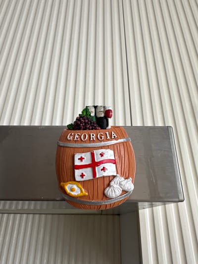 A close-up, eye-level shot captures a decorative, barrel-shaped object attached to a metallic pole, set against a background of horizontal, off-white siding. The barrel is a warm, wooden brown with silver bands. It features several embellishments: a cluster of dark purple grapes, two bottles of wine with white labels and red caps, and the Georgian flag, which is white with a large red cross and four smaller red crosses in the quadrants. The word "GEORGIA" is inscribed in white on the barrel's side. A small, golden, eye-shaped charm with a yellow center adorns the right side of the barrel. To the left of the barrel, a white, puffy embellishment resembles a bag or a cloud. The lighting is even, suggesting an overcast day or an indoor setting. The metallic pole appears to be a support structure, possibly part of a fence or a building's exterior. The scene offers no clear indication of time of day or weather conditions beyond what is suggested by the lighting. The location context of Taraz, Kazakhstan, is not visually represented in the image itself.