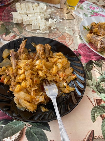 This image depicts a table laden with food and dishes in Staliyska mahala, Bulgaria. The table is covered with a patterned tablecloth featuring vintage script, botanical illustrations, and prominent red roses. In the foreground, a black, fluted dinner plate contains a partially consumed meal consisting of stewed cabbage, pieces of meat (identifiable by bones and ribs), and possibly other vegetables, all covered in a reddish-brown sauce. A silver fork, marked "STAINLESS STEEL," rests in the food. To the upper left, a clear glass plate holds numerous irregular cubes of white brined cheese. In the upper right background, a white plate with a floral rim displays further remnants of meat and stewed vegetables. Adjacent to this plate, a clear glass contains an orange beverage, and a small white cloth with pink floral accents is visible. A slice of white bread is partially visible in the far background. No individuals are present in the frame.
