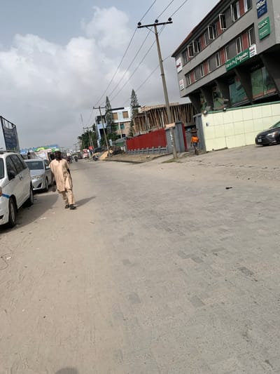 The image depicts a street scene in Jakande, Nigeria, under an overcast sky. The foreground shows a sandy shoulder transitioning into a grey interlocking paver road surface. On the left, a man wearing a light-colored tunic and trousers walks away from the camera, holding a dark object in his right hand, past parked white and silver vehicles. On the right, a multi-story building with a light-colored tiled base and red-framed windows displays business signs including "MILAN" and "Schneider Electric." A pedestrian in an orange top and dark shorts walks away from the camera along the building's base, carrying an item on their head. Further down the street, a red corrugated fence fronts a building under construction with wooden scaffolding, and utility poles with overhead power lines line the street. A black sedan is partially visible parked near the multi-story building on the far right.