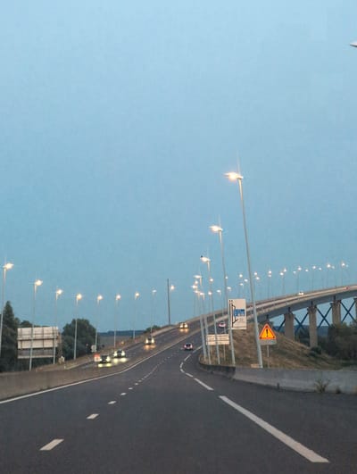The image shows a highway leading towards a large bridge, the Pont de Normandie, in Oudalle, France. The sky is a pale blue, suggesting either dawn or dusk. Numerous streetlights line the highway and the bridge, illuminating the scene. Several cars are visible on the road, their headlights on. There are road signs, including a warning sign with a yellow triangle and an exclamation mark. The bridge itself is a prominent feature, supported by large piers. Trees and vegetation are visible on the sides of the highway and below the bridge. The perspective suggests the photo was taken from a moving vehicle on the highway.
