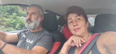 A man and a woman are seated in a car. The man, in the driver's seat on the left, is looking out the window and appears to be driving. He has a grey beard and is wearing a grey t-shirt. The woman is in the passenger seat on the right, facing the camera and smiling. She has short, dark hair and is wearing a grey tank top with a pink seatbelt. The interior of the car has black seats with red accents. Outside the window, lush green trees and foliage are visible, suggesting a rural or suburban setting. The image does not provide any specific details that would indicate a location within the city of Artvin, Turkey.
