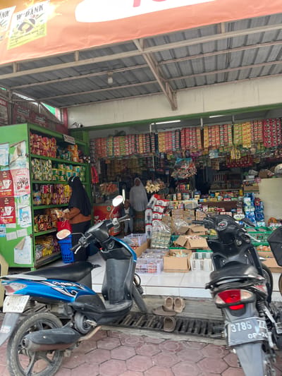 This is a street-level, eye-level, medium shot of a small grocery store in Jati Alun Alun, East Java. The storefront is open, with a corrugated metal roof supported by wooden beams. Inside, shelves are packed with various packaged goods, including snacks and beverages. A woman wearing a black hijab and an orange top is selecting items from a shelf. In front of the store, two motorcycles are parked. The one on the left is blue and black, with a license plate reading "U 5521 HAB 08-30". The one on the right is black, with a license plate reading "3786 ZZ 08-24". A small child in a white hijab and patterned skirt stands near the entrance of the store, and another adult wearing a white hijab and patterned skirt is visible inside the store. Several pairs of sandals are placed on the ground near the motorcycles. The pavement in front of the store is tiled with a brick pattern.