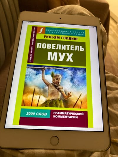 The image shows a tablet device displaying the cover of a book titled "Повелитель мух" (Lord of the Flies) by William Golding. The cover features an illustration of a young person with spears. Text on the cover indicates it is an "Эксклюзивное чтение на английском языке" (Exclusive reading in English), and also mentions "2000 слов" (2000 words) and "Грамматический комментарий" (Grammatical commentary). The tablet is resting on a person's lap, who is wearing a beige shirt. The background is out of focus and appears to be an indoor setting with soft lighting. There is no visible indication or context within the image that places the scene in Kushchovskaya, Russia.