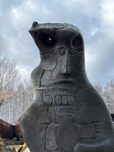 A large, dark stone sculpture, possibly a gargoyle or a stylized mythical creature, is depicted against an overcast sky. The sculpture features a prominent, elongated head with a beak-like snout and deep-set eye sockets. Its surface is intricately carved with detailed, curvilinear patterns that resemble scales, ancient hieroglyphs, or abstract designs. The texture of the stone is rough and granular, with visible etchings and some subtle cracking.
The sculpture's body, partially visible, continues the elaborate carving work, creating a sense of a richly adorned or patterned skin. The figure is angled upwards, giving it a monumental and watchful presence. The sky behind it is a mix of light gray and faint blue, indicating daytime. Bare tree branches are visible in the soft-focus background, suggesting a winter or early spring setting. Hints of architectural structures, likely made of stone or timber, can be seen in the lower right background, out of focus. No people or visible text are present in the image. The scene is located in Sukhobuzimskoye, Russia.