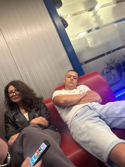 The image depicts two individuals, a woman and a man, seated on a red sofa in what appears to be an indoor waiting area or lounge. The woman, positioned on the left, has curly dark hair, wears glasses, a black leather jacket over a light-colored top, and grey sweatpants. She is looking towards the camera with a slight smile. In her lap, her hands are visible, one holding a smartphone displaying a colorful game interface. The man, on the right, is wearing a white t-shirt and light blue denim shorts. His arms are crossed, and he is looking directly at the camera with a neutral expression.
The environment features a ribbed, light-colored wall behind the woman and a blue frame structure to the right. The background beyond the blue frame is obscured by a frosted glass panel with reflective elements and what seems to be decorative signage or logos featuring a crown. The lighting suggests an indoor setting, with some artificial light sources visible, casting reflections on the glass. The overall atmosphere is casual and relaxed.
This scene does not appear to be in the city of Okop, Bulgaria, as there are no identifiable landmarks or contextual elements that would suggest this specific location.