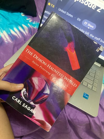The image shows a close-up, overhead view of a book titled "THE DEMON-HAUNTED WORLD" by CARL SAGAN, lying on top of a laptop. The book is open, revealing its cover which features a dark, abstract background with a bright red object and a crystal ball reflecting colorful light. A person's hand is holding the book. The laptop is silver with an "Intel Core i3" sticker on it. Another blue-covered item is partially visible underneath the book, showing text like "Episode 2", "United States", "EN", and a "Share" button. The scene is set on a purple, textured surface, possibly a bed or blanket. The lighting suggests it could be indoors, during the day or evening. The overall impression is one of casual study or leisure.
