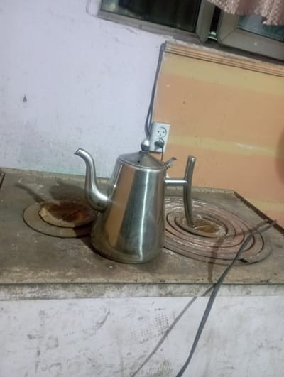 A shiny metal teapot with a curved handle and spout is positioned on a rustic, circular heating element. The heating element appears to be part of an older stove, possibly a wood-burning stove, with concentric rings etched into its surface. The teapot is plugged into a white electrical outlet mounted on a peach-colored wall. The wall is textured, and the outlet has two sockets. To the left of the outlet, a window with a reflective surface and a curtain is partially visible. The background wall is a lighter, rough-textured color, showing signs of age and wear, with faint smudges and marks. A dark electrical cord snakes from the teapot towards the right side of the frame. The lighting suggests an indoor setting, likely during the day. The overall scene conveys a sense of utilitarianism and perhaps a humble domestic environment. There is no visible text in the image.