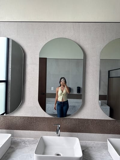 A woman is reflected in a bathroom mirror. She is wearing a light-colored tank top and blue jeans, holding a cell phone. The bathroom has three mirrors and two white square sinks. The mirrors and sinks are mounted on a wall with gray tile. There is a strip of brown mosaic tile separating the mirrors from the sinks. The location is Santa Ana Yancuitlalpan (Chilhuacán), Mexico.