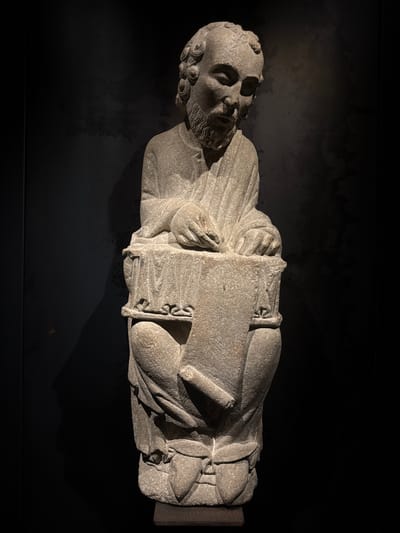A stone sculpture of a man is displayed against a dark background. The man is seated with his legs crossed and appears to be writing on a scroll that rests on a stone block in front of him. He has a beard and curly hair, and wears a draped robe. The sculpture is illuminated from the side, casting a shadow on the dark wall behind it. This sculpture is the “Maestro Mateo,” a master sculptor, and is located in the Museo de las Peregrinaciones de Santiago in Santiago de Compostela, Spain.