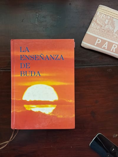 A close-up shot presents a book titled "LA ENSEÑANZA DE BUDA" on a dark wooden surface. The book's cover features a sunset-like image. In the upper left corner, part of a beige wallet with "PARIS" printed on it is visible. A black remote control sits on the right side of the frame, also on the wooden surface. The scene conveys a sense of reading or relaxation.