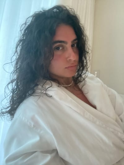 A woman with long, dark, curly hair is lying in bed. She is partially covered with a white blanket or sheet. She is wearing a pearl necklace. The location is indoors, possibly a bedroom, but not conclusively the city of El Caracol, Mexico.