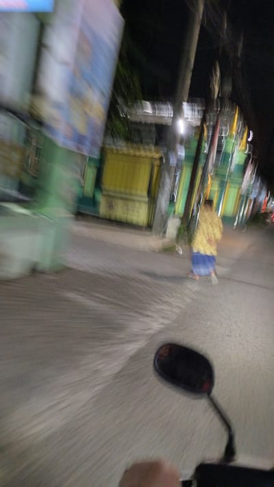 This is a motion-blurred image taken at night from the perspective of someone riding a motorcycle in Bangsri, Indonesia. In the mid-ground, a person is walking away from the camera, wearing a yellow patterned garment over a blue sarong. The background shows a street scene with buildings that appear to be shops or residences, featuring green and yellow facades. A large, blurry blue and white sign is visible on the left side of the image, and a motorcycle mirror is in the foreground. The road surface is paved with what appears to be interlocking bricks or cobblestones.