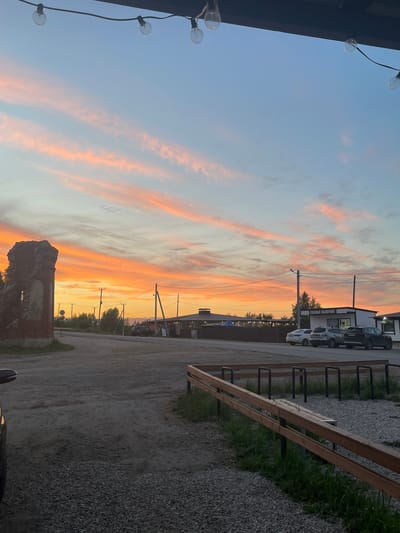 The image depicts a roadside scene in Shiryaevo, Russia, during a sunset. The sky is filled with streaks of orange and pink clouds, transitioning to a pale blue at the top. String lights are suspended overhead.
In the foreground, a gravel and dirt area is visible, with a wooden bench and bike racks. To the left, a partially ruined brick structure stands prominently. A dirt road leads into the distance, flanked by trees and utility poles.
Further down the road, several buildings are visible, including one with a sign that appears to read "СВЕЖАЯ ВЫПЕЧКА" (Fresh Pastry). Three cars are parked in front of the buildings: a white sedan, a silver SUV, and a black SUV. The overall impression is one of a rural or semi-urban setting at dusk.