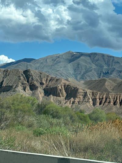 The image depicts a mountainous landscape in Kok Moynok Eki, Kyrgyzstan, featuring rugged terrain, sparse vegetation, and a partly cloudy sky. The scene appears to be taken from a roadside, with a paved road visible along the right edge. The mountains exhibit varying shades of gray and brown, indicating different rock types or levels of weathering. Sparse patches of greenery suggest limited vegetation growth in the area. The sky is partly covered with clouds, providing a mix of sunlight and shade.