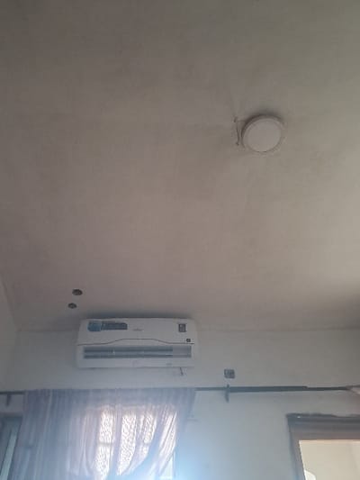 An interior view in Langbasa, Nigeria, depicts a room with an off-white ceiling and wall. A white, horizontal split-unit air conditioner is mounted on the wall below the ceiling, slightly to the left of the frame's center. Above the air conditioner, two small, dark circular holes are present in the ceiling. To the upper right, a round, white, surface-mounted light fixture is attached to the ceiling. A sheer, light-colored curtain covers a window on the lower left, illuminated by external light. A dark curtain rod supports the curtain. A dark-colored electrical conduit runs horizontally along the wall above the curtain rod, with a square electrical component attached to it. A wooden frame, possibly for a window or door, is partially visible on the far right. No individuals or active interactions are present.