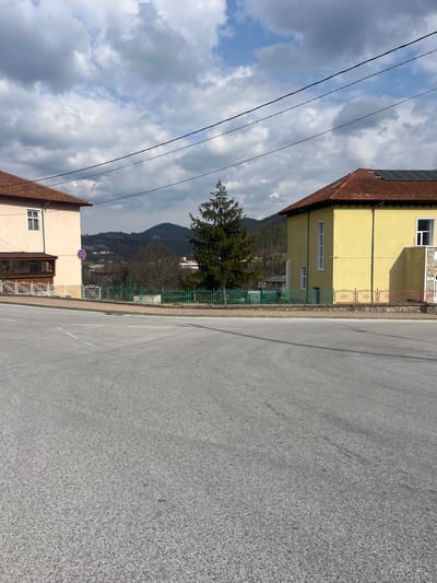 The image displays an outdoor street scene in Srednogortsi, Bulgaria, under a partially cloudy sky. The foreground is dominated by a gray asphalt road, featuring faint white dashed line markings. No individuals or vehicles are visible.
To the left, a two-story building with a light beige exterior and a brown tiled roof is present. It has white-framed windows and a ground-level enclosed wooden and glass extension. A circular "No Stopping" sign (red circle with a diagonal line on a blue background) is mounted on a pole near this structure. A white metal fence with decorative elements separates the property from the paved sidewalk.
To the right, a two-story building with a yellow facade and a brown tiled roof is visible. The roof features several solar panels. This building also has white-framed windows and a partially visible white vertical architectural element on its side. A multi-colored fence, consisting of green, red, and white sections with decorative patterns, borders this property.
Centrally, between the two buildings and set back from the road, a tall, conical evergreen tree stands. Behind it, a green metal fence and a white basketball hoop structure are discernible. Three black overhead power lines stretch horizontally across the upper portion of the image. The background reveals rolling, tree-covered mountains with scattered distant structures, under a sky with both blue patches and substantial white and gray clouds. The overall scene appears devoid of human activity.