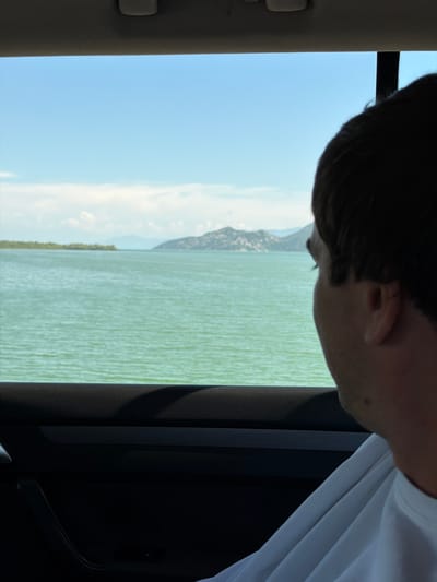 The image, taken from inside a vehicle, captures a view of a wide body of water with distant hills and a bright blue sky. The water is a vibrant green, with small ripples suggesting a gentle breeze. A person's head and shoulder, partially obscured by the frame, are visible in the foreground, looking out the window. The interior of the car is visible at the bottom, with dark-colored upholstery and a door handle. The scene appears to be located in Kruševica, Montenegro, given the characteristic landscape of the region.