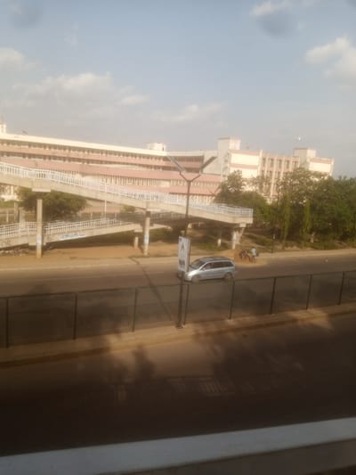 The image shows a pedestrian overpass spanning across a road in Unguwan Dosa, Nigeria. A silver minivan is driving on the road beneath the overpass. In the background, a large, multi-story building with light-colored walls and rows of windows is visible. Lush green trees surround the building and the overpass. The sky is partly cloudy, suggesting it is daytime. The scene appears to be a busy urban environment with infrastructure for both pedestrians and vehicles. There is some graffiti on the supports of the overpass. A tall pole with a sign is visible on the left side of the overpass. There are a few people visible near the road, possibly crossing or observing the traffic.