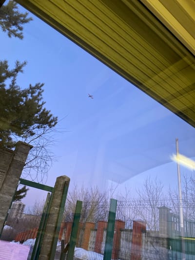 The image presents an outdoor scene in Марфино, Russia, viewed through a window. A reflection of horizontal yellow-green blinds occupies the upper margin of the frame. The sky is clear and light blue, featuring a small, reddish-orange airplane traversing from left to right in the upper-mid section. To the left, a portion of a dark green coniferous tree is visible. Below it, and extending across the mid-ground, are numerous bare deciduous trees and shrubs, indicating a winter or early spring season. Patches of snow cover the ground intermittently. Various fencing structures are present: a large, weathered concrete pillar stands on the left. Adjacent to it, a green metal gate or fence with concrete posts is visible. Further back, a chain-link fence is discernible. In the mid-ground, a multi-sectioned fence features alternating reddish-brown and gray concrete elements with wire mesh panels. In the distant background, a tall, light-colored residential or commercial building is partially observed through the bare vegetation. No individuals are depicted in the scene. The ambient lighting suggests daylight.