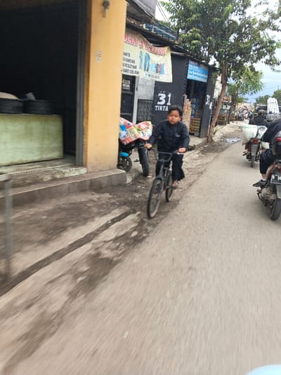 A young boy on a dark bicycle is captured in motion on a paved road in Cangkuang Kulon, Indonesia. He is wearing dark clothing and no footwear, pedaling towards the right side of the frame while looking forward. The road surface exhibits signs of wear, including cracks and wet patches, suggesting recent moisture. To the immediate right of the boy, a motorcycle is partially visible, also in motion, with a rider wearing dark attire. Further down the road, additional motorcycles and riders are present.

On the left side of the frame, a light yellow/orange building with a raised concrete sidewalk is visible. An open doorway leads into a dark interior where dark circular objects and a light green rectangular object are discernible. Attached above an adjacent dark-colored structure is a white banner with Indonesian text, advertising "JAHIT & SABLON" (Sewing & Screen Printing) services, including "WANITA" (women's), "KANCING & RESLETING" (buttons & zippers), and "KAOS" (T-shirts), along with a phone number. Below the banner, the number "31" and the word "TIRTA" are displayed. A blue scooter is parked near this structure, covered with a patterned fabric over its seat. A small tree with green foliage stands between the buildings and the road. The sky above is overcast. The foreground exhibits motion blur, indicating the photograph was taken from a moving perspective.