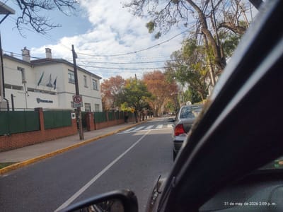 Captured from inside a car, the image presents a street scene in Martínez, Argentina, on May 31, 2026, at 2:09 p.m. A silver Ford Focus is parked ahead on the right side of the road. On the left side of the street, a building with white walls and multiple flags flying stands behind a green fence atop a brick base. A "No Parking" sign is visible near the building. The road is paved and marked with a white dividing line. Crosswalk stripes appear further down the road. Trees line the street, some displaying autumn foliage. A pedestrian is walking along the sidewalk. The sky is partly cloudy. The interior of the car from which the picture is taken obscures a portion of the right side of the view.