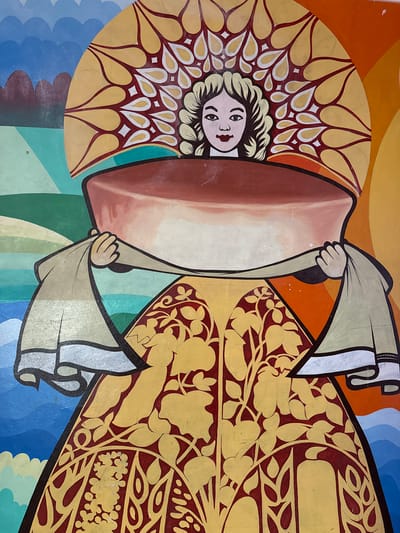 The image is a close-up of a vibrant, stylized mural depicting a person, likely a woman, with light-colored hair and distinct facial features. The figure is adorned in flowing garments that are predominantly light brown and yellow, embellished with intricate floral and leaf patterns in dark red and yellow against a yellow background. The figure's face is centrally positioned, looking directly at the viewer with wide eyes and red lips.

The figure appears to be holding a large, soft object that is a gradient of light brown and white. The surrounding colors are bold and abstract, featuring swathes of orange, blue, green, and lighter blue in curved, wavy patterns. The artwork has a distinct, almost folk-art style, with bold outlines and flat areas of color.

There are no visible signs of a specific time of day, weather conditions, or explicit location cues within the mural itself, other than the context provided of "Лащ-Таяба, Russia." No text is visible in the image. The overall impression is of a colorful and symbolic representation, possibly part of a larger narrative or artistic installation.