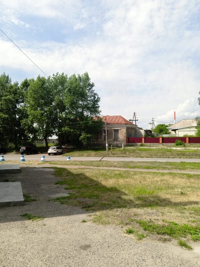 A street view in Pavlovsk, Russia, on a partly cloudy day. In the foreground, a paved area with some patchy grass and a shadow from a building. Beyond this, a road with two white and blue traffic cones. On the other side of the road, a grassy area with a red metal fence. Behind the fence, a light brown building with a red roof, and to its right, another building with a light-colored roof and a tall red and white chimney. Several large trees with green foliage are present. Two cars are parked on the side of the road near the trees.