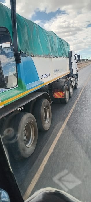 A truck's rear tires and a portion of its tarp-covered cargo are seen on a paved road with a yellow line. The truck's side has a blue and white design with lettering. A red and yellow striped safety marker is visible near the tires. The sky is cloudy. The image is taken from a higher perspective, likely from another vehicle.