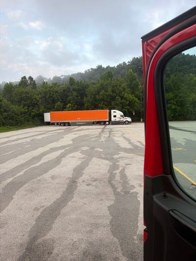 A red truck cab door is open on the right side of the frame, obscuring the foreground. In the middle ground, a white semi-truck with an orange trailer is parked in a lot. The trailer has "schneiderjobs.com" printed on its side. The lot is paved and appears wet, with dark, elongated streaks across the surface. In the background, a dense line of green trees covers a hillside under a cloudy sky. The scene appears to be in a rural or semi-rural area. The exact location in Wildwood, United States is not discernible from the image alone.