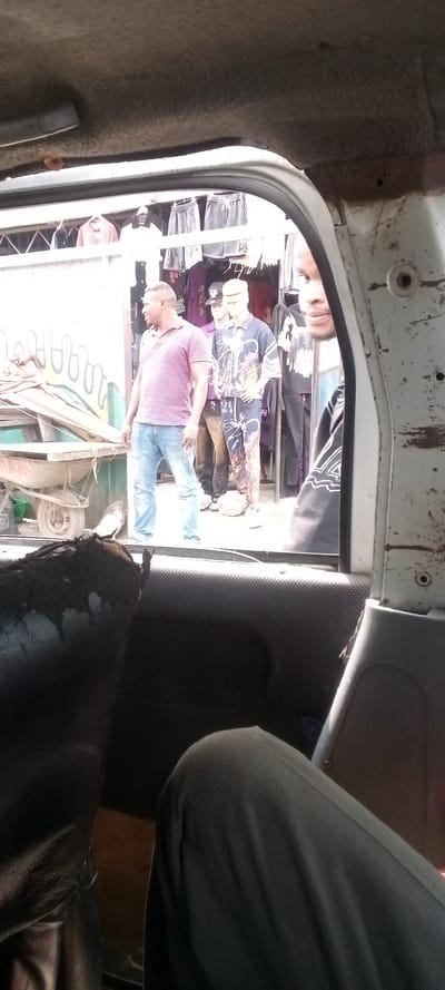 The image shows the interior of a vehicle with a person sitting in the front passenger seat, looking out the window. The window looks out onto a street scene in Igrita, Nigeria (latitude: 4.957, longitude: 7.012). The person in the vehicle is wearing dark-colored clothing, and their legs are visible in the shot. They are looking at a man in a pink shirt and jeans standing on the sidewalk, looking back at them. Behind the man in the pink shirt, there is a shop with mannequins dressed in colorful clothing. The shop has a white awning and a colorful mural painted on the wall. The vehicle is a white van with a dirty interior. The image is slightly blurry and the lighting is poor.