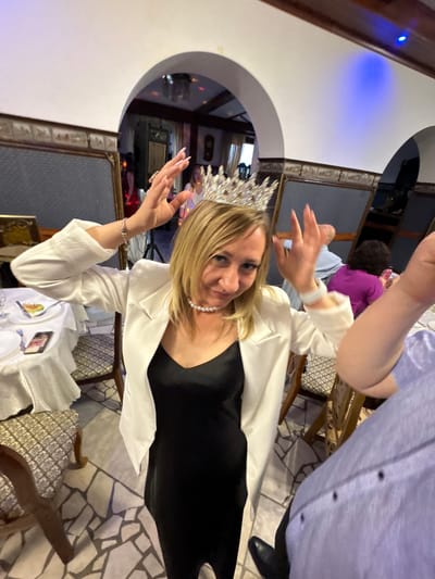 A woman is wearing a tiara and a pearl necklace, and a white blazer over a black dress. She is inside what appears to be a restaurant, possibly in Zavod Mikhailovskii, Russia, based on the user's prompt. The floor is tiled with irregular stone pieces. In the background, there are arches, decorated ceilings with lights, and other patrons at tables. One person's arm and shoulder, wearing a blue shirt, are prominently in the foreground, obscuring part of the woman and the scene. The woman is holding her hands up, near her head, and appears to be posing for the photo. There are tables with white tablecloths set for dining.